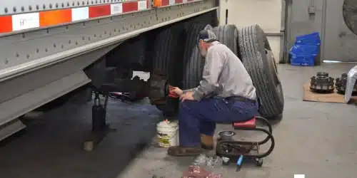 A truck service technician completes a trailer inspection, checking tire tread and pressure while kneeling by a tire.