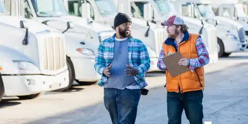 Two men walk in front of a fleet of white semi trucks with a clipboard having a conversation about fleet management solutions.